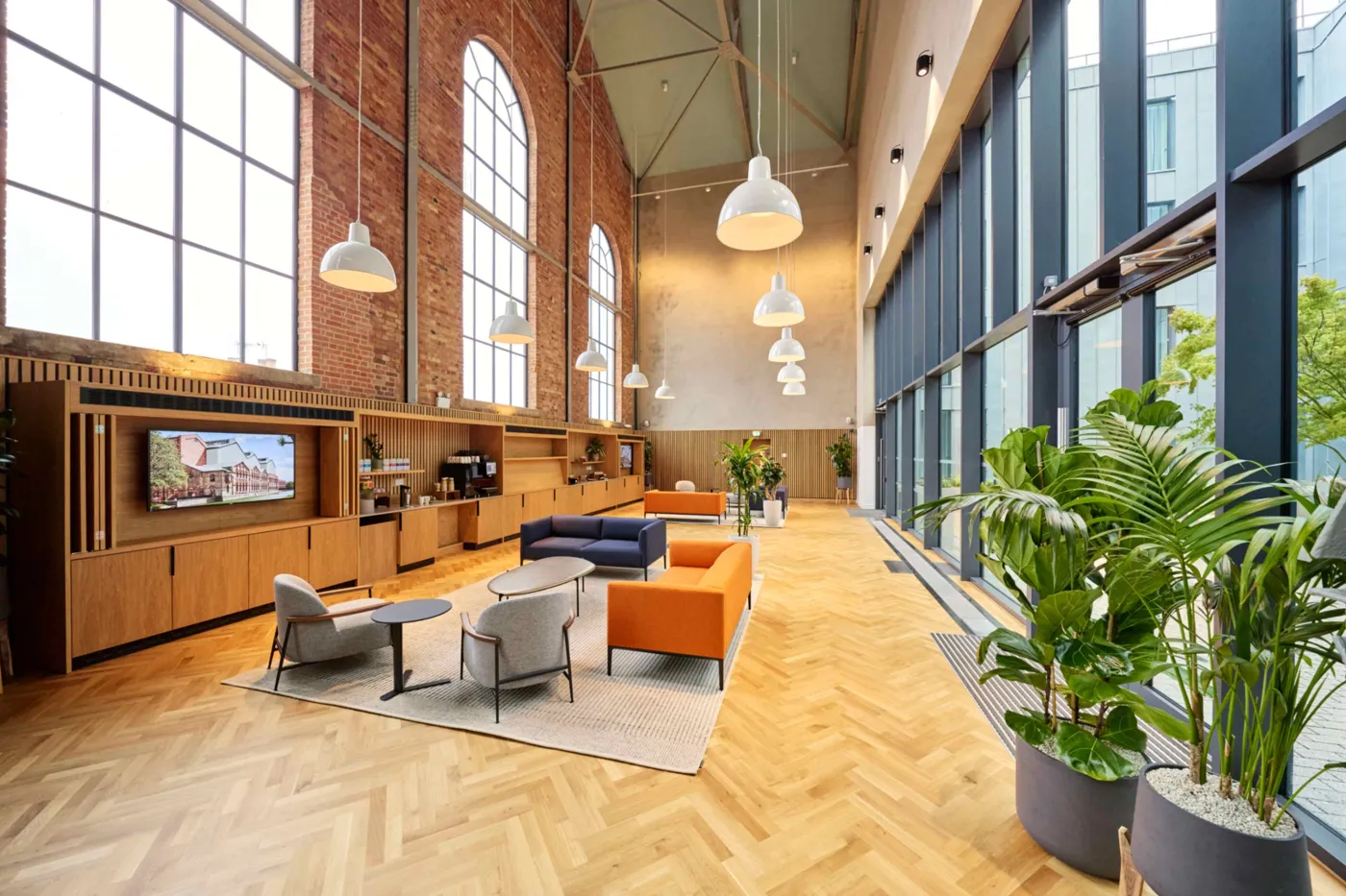 A long lounge with orange armchairs, plants, wooden shelving and high arched windows in a brick wall