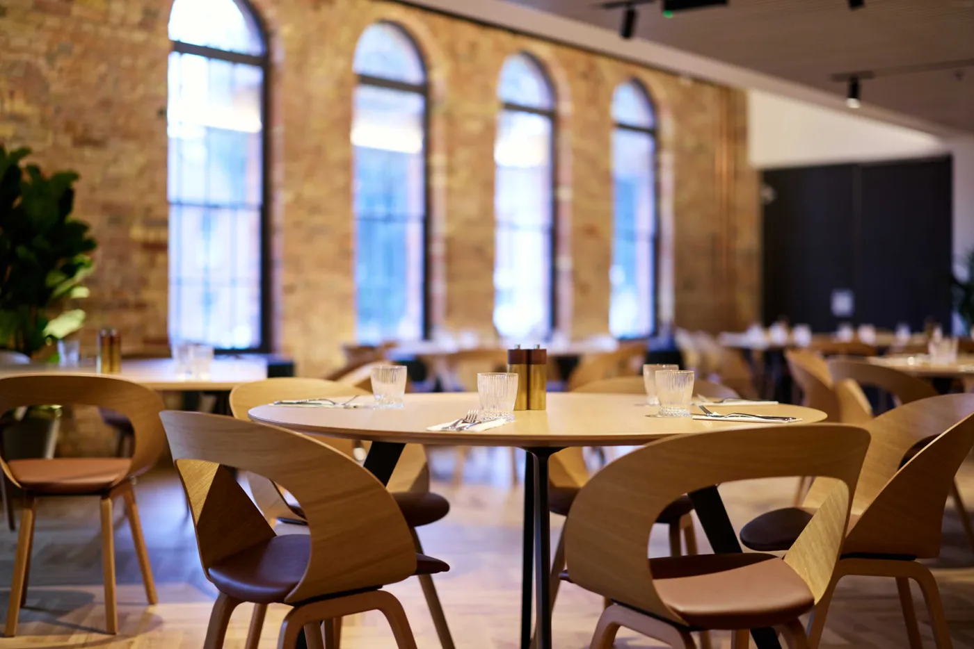 A dining room with round tables and wooden chairs beneath arched windows in a brick wall
