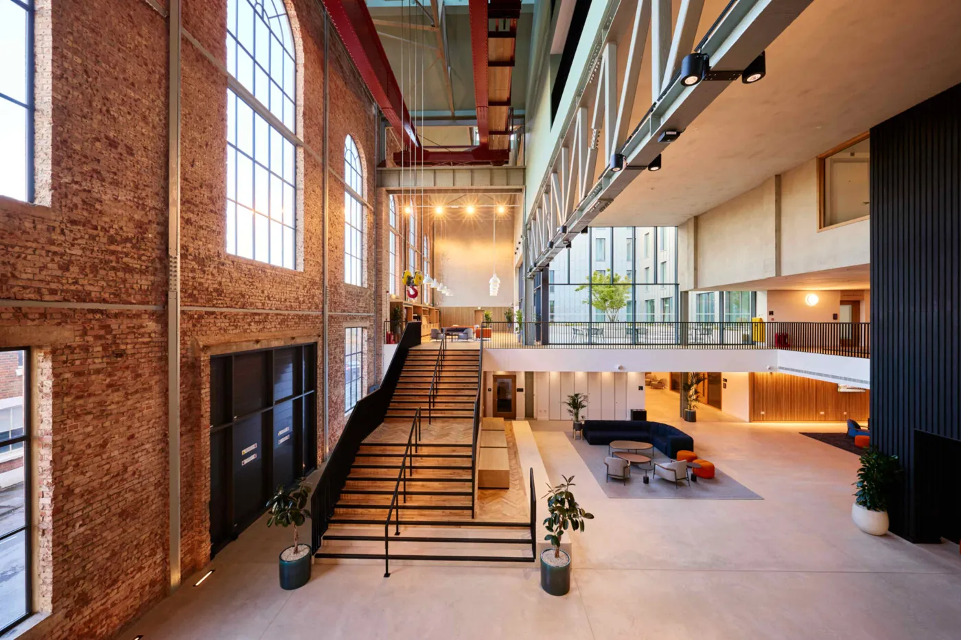 A tall interior hall with exposed brick walls, arched windows, a wide staircase and red steel beams overhead