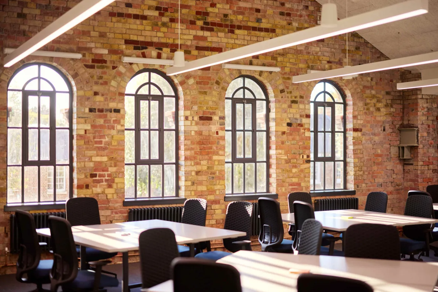 A classroom with desks and black chairs in front of a brick wall with four arched windows